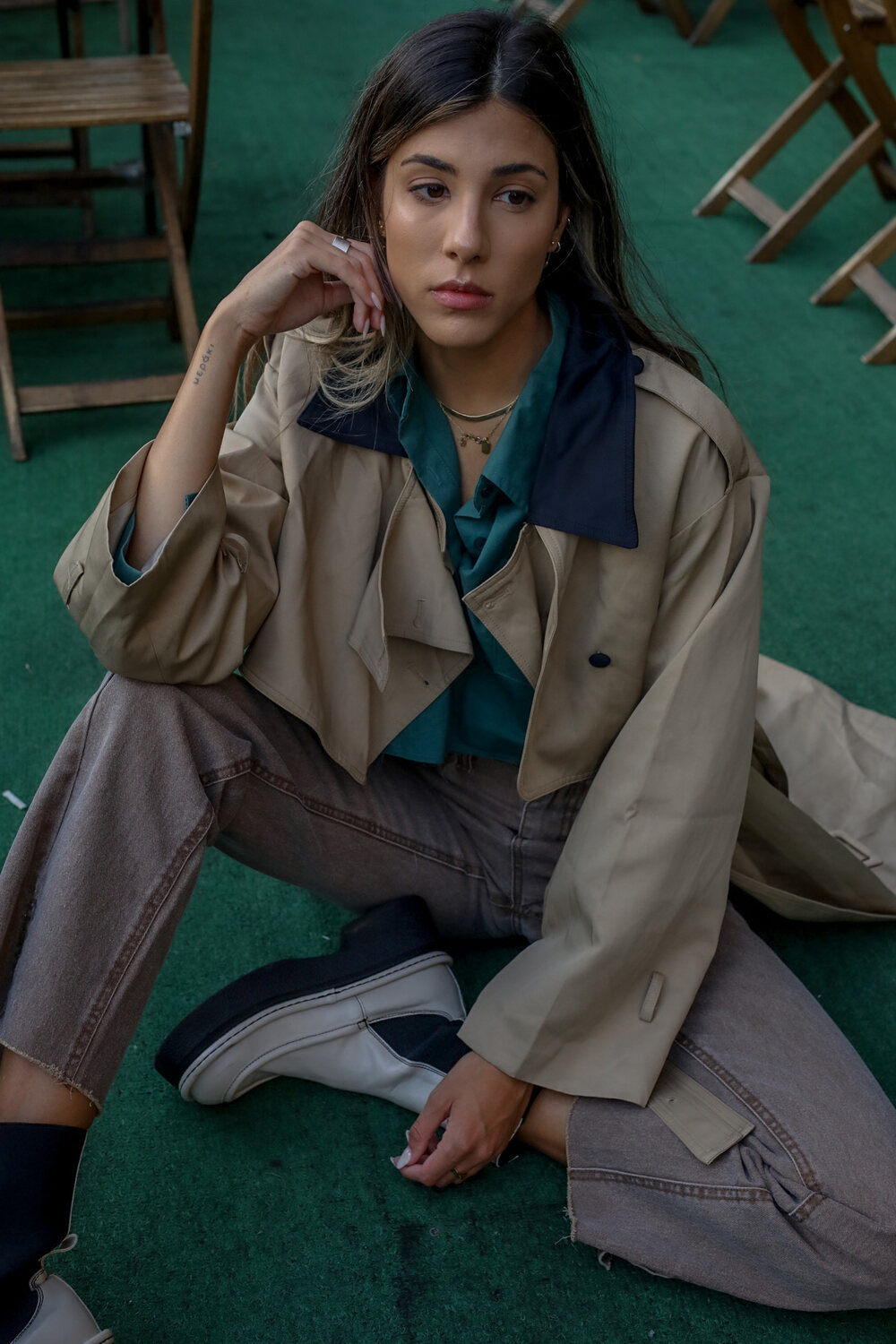 Model in a layered trench coat seated against a deep green backdrop