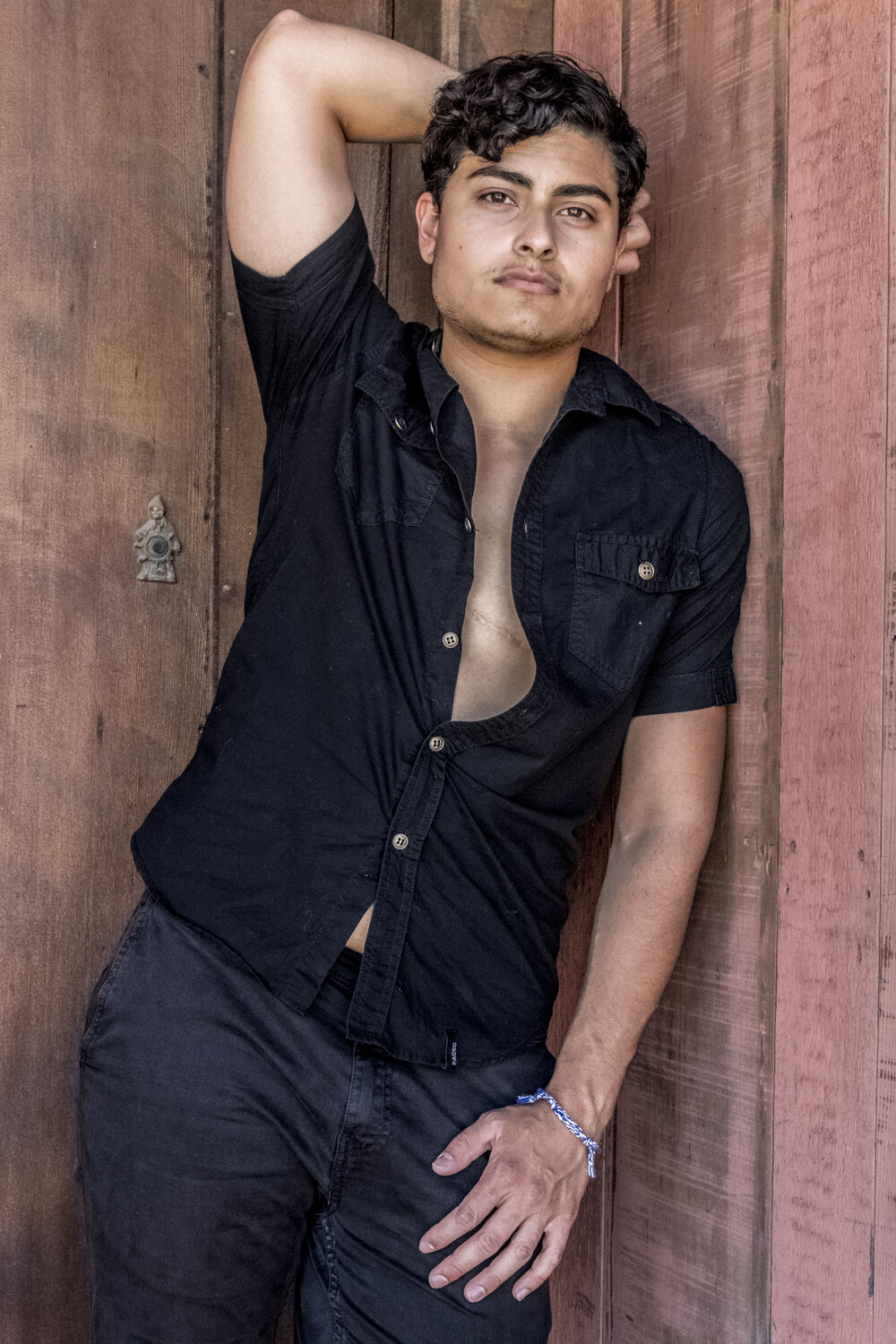 Man in an open black shirt leaning against a weathered wooden wall