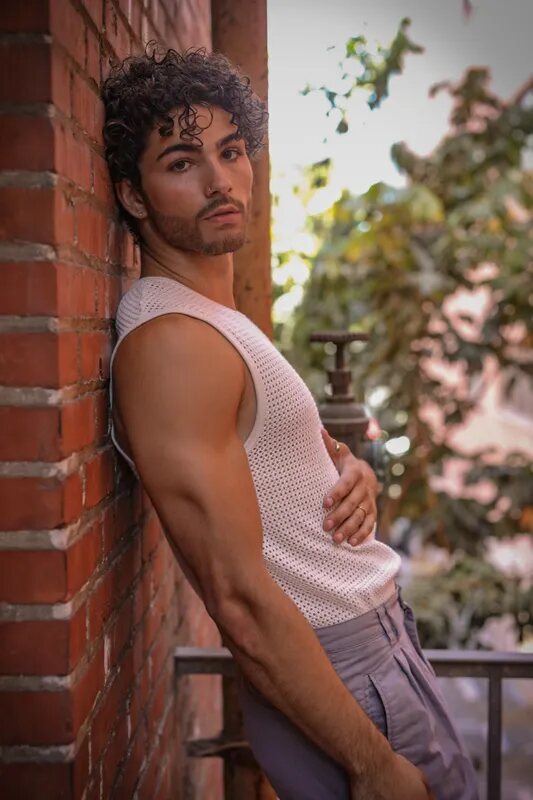Man in a blush tank top leaning against a brick wall with greenery